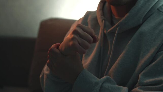Young man in dim room suffering from hand pain, massaging wrist, clenching fist, and doing circular hand movements to relieve pain. Concept of injury, strain, recovery, muscle tension, and self treatm