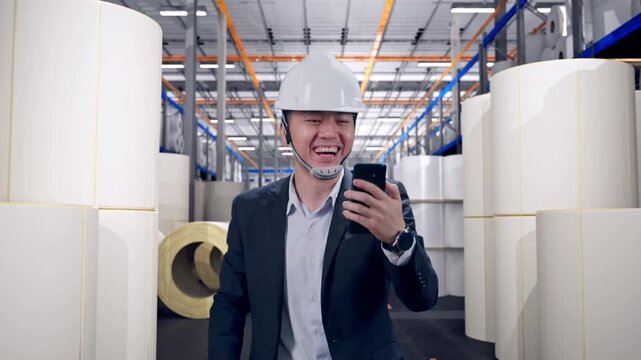 Front View of Asian Businessman Use Smartphone, Video Call While Walking at Warehouse with Rows of Stacked White Polymer and Paper Rolls