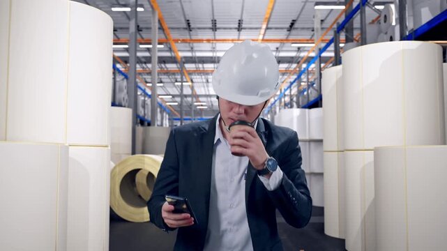 Front View of Asian Businessman Use Smartphone, Drink Coffee While Walking at Warehouse with Rows of Stacked White Polymer and Paper Rolls