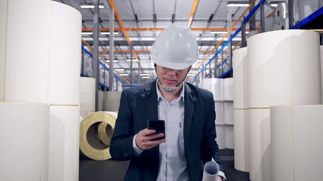 Front View of Asian Businessman Use Smartphone, Holding Blueprint While Walking at Warehouse with Rows of Stacked White Polymer and Paper Rolls