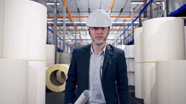 Front View of Asian Businessman Holding Blueprint While Walking at Warehouse with Rows of Stacked White Polymer and Paper Rolls