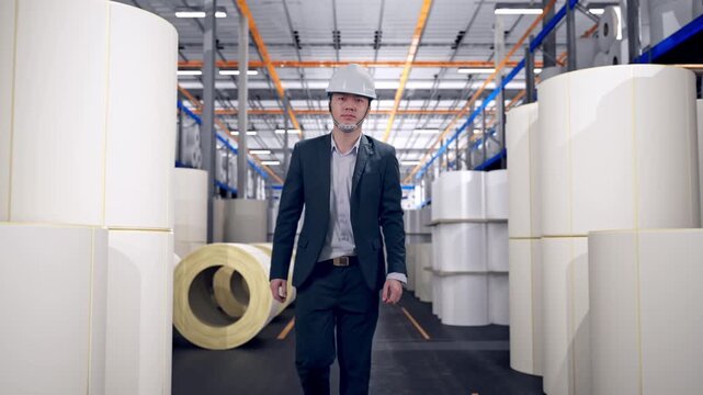 Front View of Asian Businessman Walking at Warehouse with Rows of Stacked White Polymer and Paper Rolls