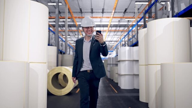 Front View of Asian Businessman Use Smartphone, Video Call While Walking at Warehouse with Rows of Stacked White Polymer and Paper Rolls