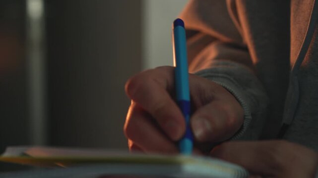 Person making notes and writing ideas in a notebook during evening time. Focus on planning, studying, brainstorming, and creative thinking in a home environment. Close-up shot with soft light.