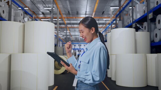 Side View Of Asian Female With Her Tablet at Warehouse with Rows of Stacked White Polymer and Paper Rolls, She Raises Her Fist Up With Screaming Goal After Check On The Tablet