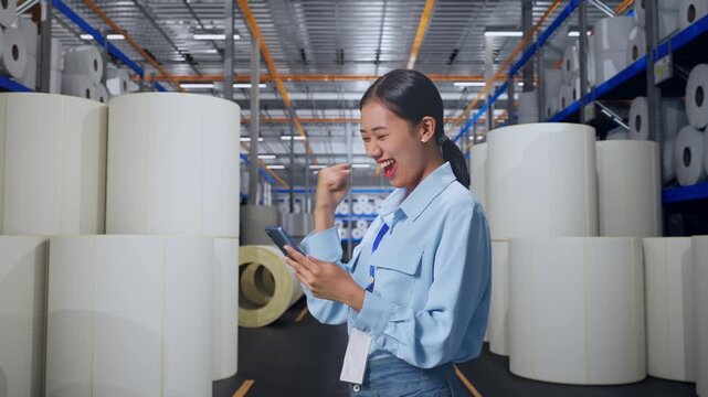 Asian Female Professional Worker With Her Smartphone at Warehouse with Rows of Stacked White Polymer and Paper Rolls, She Raises Her Fist Up With Screaming Goal After Check On The Smartphone