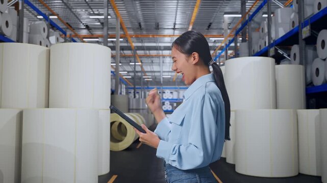 Side View Of Asian Female With Her Tablet at Warehouse with Rows of Stacked White Polymer and Paper Rolls, She Raises Her Fist Up With Screaming Goal After Check On The Tablet