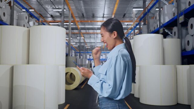 Asian Female Professional Worker With Her Smartphone at Warehouse with Rows of Stacked White Polymer and Paper Rolls, She Raises Her Fist Up With Screaming Goal After Check On The Smartphone