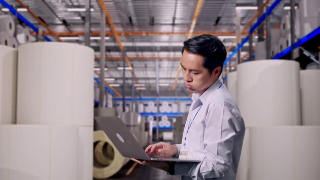 Asian Male Worker Use Laptop at Warehouse with Rows of Stacked White Polymer and Paper Rolls,  He Is Nodding His Head With Dissapionted