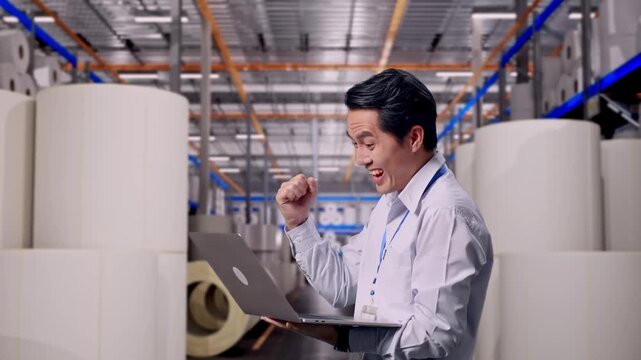 Asian Male Worker Standing With His Laptop at Warehouse with Rows of Stacked White Polymer and Paper Rolls, He Raises His Fist Up With Screaming Goal After Typed On The Laptop