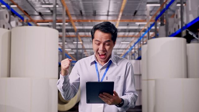 Asian Male Worker Standing With His Tablet at Warehouse with Rows of Stacked White Polymer and Paper Rolls, He Raises His Fist Up With Screaming Goal