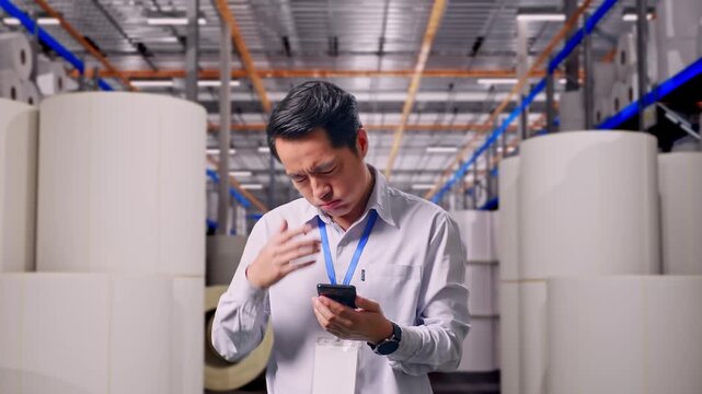 Asian Male Worker Standing With His Smartphone at Warehouse with Rows of Stacked White Polymer and Paper Rolls, He Is Nodding His Head With Dissapionted