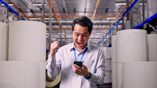 Asian Male Worker Standing With His Smartphone at Warehouse with Rows of Stacked White Polymer and Paper Rolls, He Raises His Fist Up With Screaming Goal