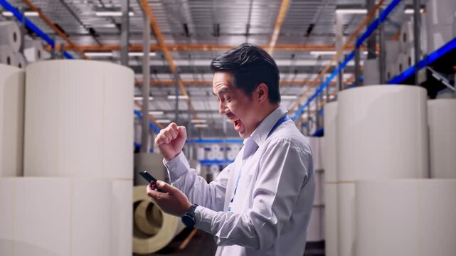 Asian Male Worker With His Smartphone at Warehouse with Rows of Stacked White Polymer and Paper Rolls, He Raises His Fist Up With Screaming Goal After Check On The Smartphone