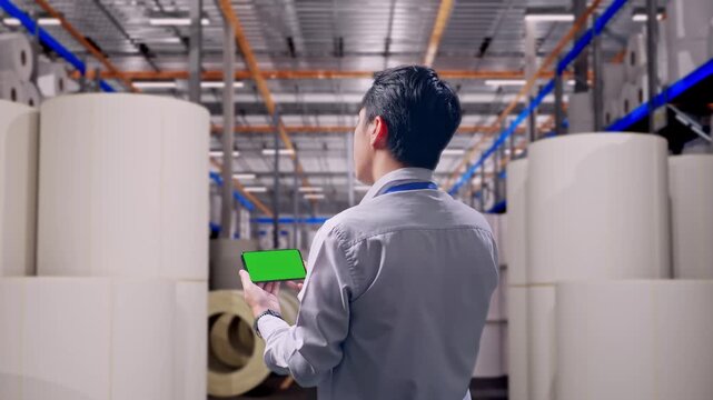 Back View Of An Asian Male Worker Standing at Warehouse with Rows of Stacked White Polymer and Paper Rolls, Checking On Smartphone With Green Screen And Look At The Factory Once