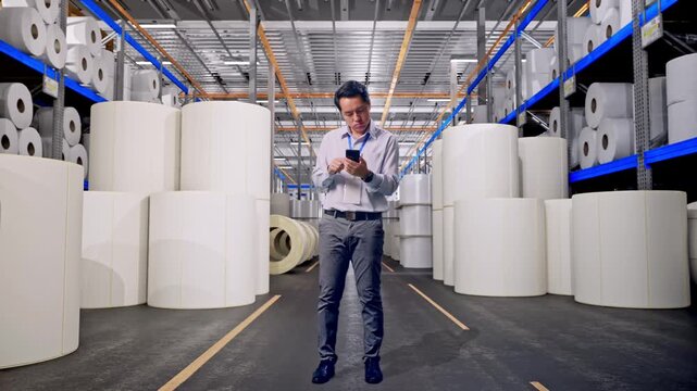 Full Body Of Asian Male Worker With His Smartphone at Warehouse with Rows of Stacked White Polymer and Paper Rolls, He Is Nodding His Head With Dissapionted