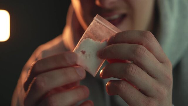 Close-up of a man in a dimly lit environment holding a small transparent zip bag containing white powder, possibly cocaine. The image represents themes of drug addiction, abuse substance, illegal drug