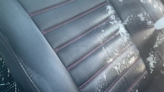 A detailed horizontal shot of a hand spraying cleaning product onto a gray fabric car seat. The image captures fine mist settling on the textured textile surface. High quality FullHD footage