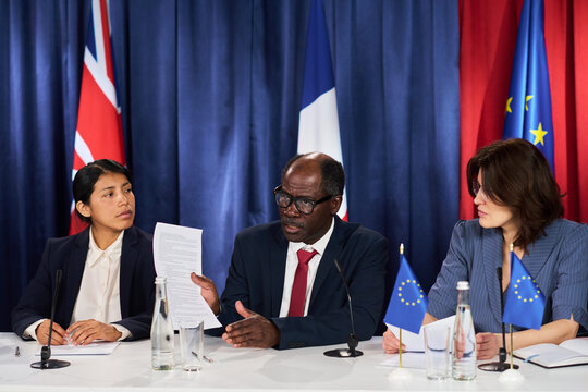 Medium shot of diverse business leaders reviewing document during international press conference, discussing policy alignment. Ideal for diplomacy, government relations