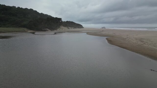 A coastal lagoon sits adjacent to the open ocean, separated by a narrow land barrier. Surrounding hillsides are densely vegetated, and cloud cover is visible across the sky.