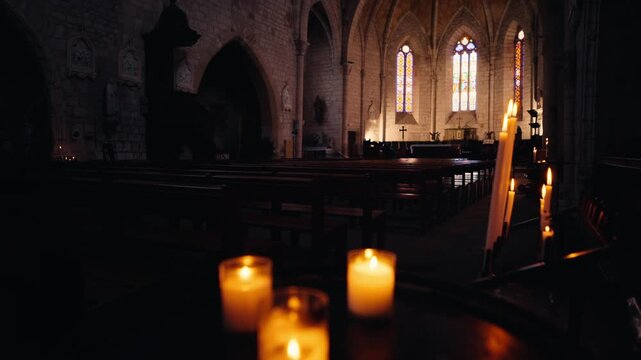 Row of burning candles in a dark church interior in Monpazier, Dordogne, with wooden pews and gothic stained glass. Slow motion tracking shot.