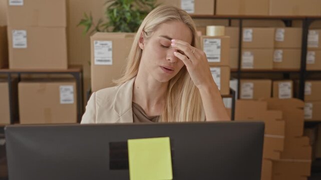 Woman rubbing eye at computer desk surrounded by parcel boxes and sticky note on monitor in a storage building; stress deadline.