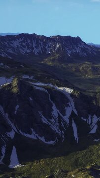 The scene shows mountains with rocky slopes and valleys under a clear sky. Snow is visible on some peaks. The landscape appears vast and open. Light touches the terrain, revealing details.