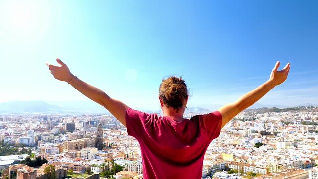 Young man with arms outstretched celebrating over Malaga city panorama, Spain. Success, freedom and travel concept with urban landscape and blue sky in Andalusia.