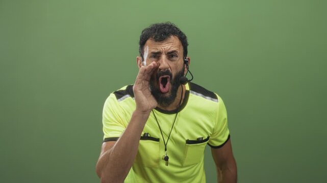 Man in neon yellow referee uniform with whistle on lanyard, hand to mouth shouting into headset microphone in studio; urgent command.
