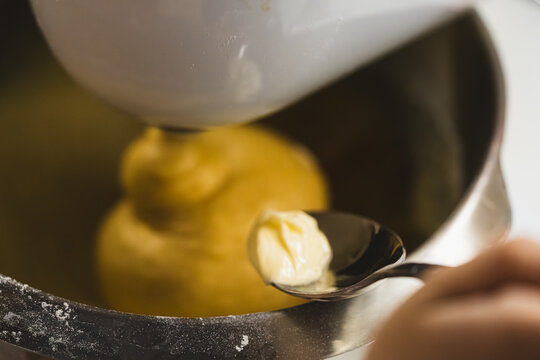 Series of photos showing the process of adding soft butter into dough. Dough in a planetary mixer metal bowl. A tablespoon with butter held above the bowl. Home cooking. 
