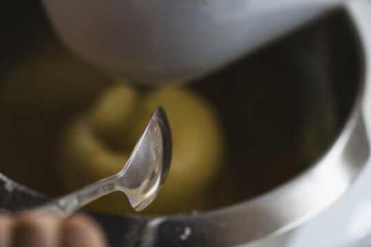 Series of photos showing the process of adding soft butter into dough. Dough in a planetary mixer metal bowl. A tablespoon with butter held above the bowl. Home cooking. 