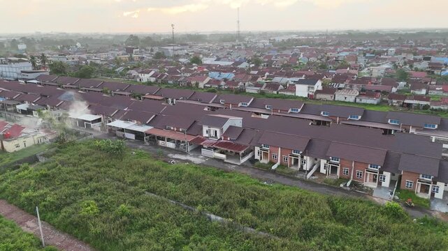 A drone shot reveals a suburban neighborhood at sunset, with rows of dark-roofed houses bordering misty green fields and distant settlements under a soft sky.