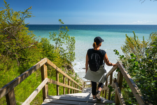 A woman with a backpack descends wooden stairs through green beech trees (Fagus sylvatica) toward the milky, turquoise Baltic Sea and clear sky. M&oslash;ns Klint, Denmark.