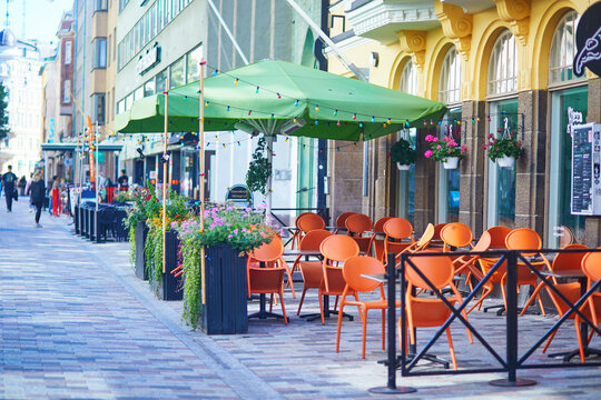 Charming outdoor cafe terrace with orange chairs, green umbrellas and flower planters along a pedestrian street in Helsinki, Finland, creating a lively urban summer atmosphere