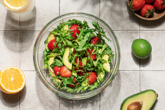 Fresh summer salad with avocado arugula and strawberries in glass bowl on gray tile table, top view healthy gourmet dish
