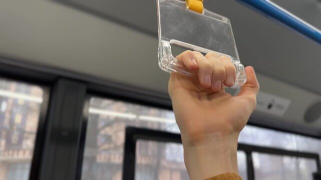 A hand grips a bus handrail. The hand is holding a clear plastic holder attached to a blue pole in a public transport setting.
