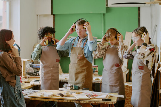 Teenagers adjusting protective earmuffs during woodworking class, preparing for practical training with instructor. Useful for education, vocational skills, school workshop promotion