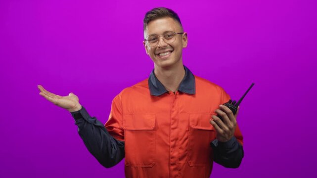 Young mechanic man holding walkie radio in right hand with left hand palm up, smiling in a studio with purple backdrop; friendly.