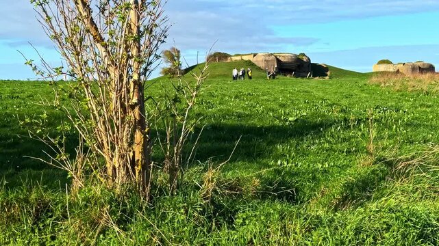 Wide shot of historic German artillery battery casemate at Longues-sur-Mer with tourists across green coastal field under blue sky in Normandy France