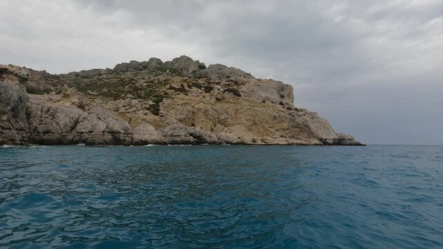 Stunning view from the water surface of rugged limestone cliffs and rocky headland in Souda Bay, Chania, Crete, Greece. Deep turquoise Mediterranean Sea under dramatic overcast sky.