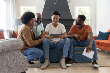 Obraz premium Diverse male friends leaning on blue ottoman in living room watching tablet holding notebook bottle
