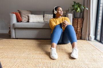 Obraz premium African American woman sitting on woven rug in living room wearing mustard knit headphones