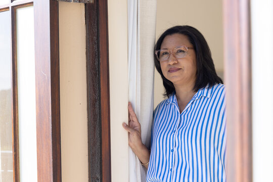 Mature Asian woman standing at doorway in striped shirt, touching curtain, looking out, copy space