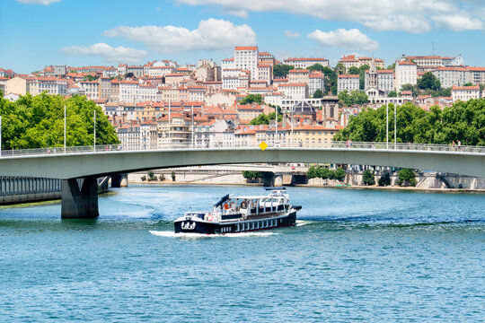 A sightseeing boat cruises along the Sa&ocirc;ne River in Lyon, passing beneath the modern Pont Juin with the historic, sun-drenched buildings of the Croix-Rousse hillside rising steeply in the background.