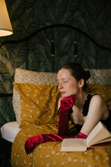 Fototapeta premium Elegant woman with frecklesin black dress and red gloves reading book in vintage interior with warm light.