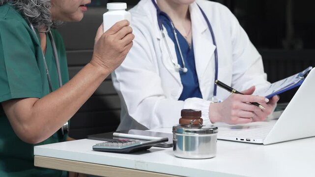 Doctor consulting with patient holding medication bottle in clinic office, with laptop, clipboard, calculator and pill container on desk, healthcare and prescription discussion.