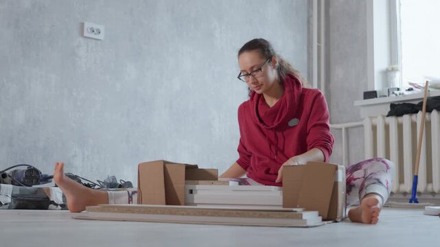 Person Assembling Furniture Calmly. Woman Quietly Putting Together Flatpack Shelf With Tools Nearby. Woman Seats On Ground Assembling Flatpack Shelving Unit With Tools And Instructions Around