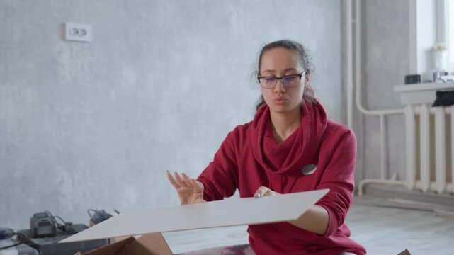Caucasian Woman Assembling Furniture In Empty Apartment, Sitting On Floor Unpacking White Panels From Cardboard Box, Focused Expression, Radiator And Tools In Background, Natural Light, DIY Flatpack
