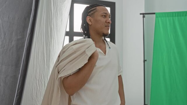 Man holds jacket over shoulder in studio with green screen and window light, facing right with braided hair and standing pose; confidence.