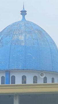 A vertical tilt-down showing a bright blue mosque dome and its unfinished lower levels. The exterior features arched windows, concrete pillars, and construction materials.